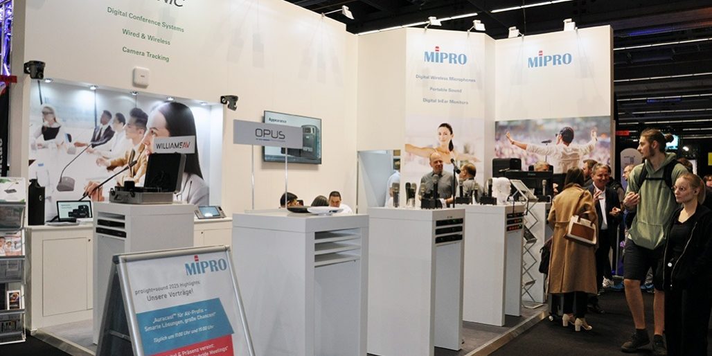 MIPRO Prolight + Sound 2025 Der Messestand von MIPRO auf der Prolight + Sound 2025 präsentiert sich hell, offen und professionell. Im Zentrum stehen mehrere Produktinseln mit digitaler Drahtlostechnik – darunter Mikrofone, Taschensender und In-Ear-Systeme. Ein aufgestelltes Rollup kündigt spannende Vorträge zu digitalen Mikrofonsystemen, smarter Bühnenpraxis und erfolgreicher Medientechnik an. Die große Besucherzahl am Stand sowie das rege Interesse an den präsentierten Lösungen unterstreichen die Relevanz von MIPRO im professionellen Audiomarkt. Begleitet wird die Ausstellung von auffälligen Visuals, die die Einsatzbereiche der Technik – von Business bis Bühne – eindrucksvoll in Szene setzen.