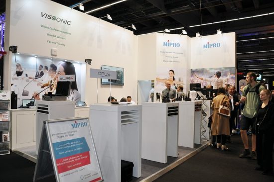 Der Messestand von MIPRO auf der Prolight + Sound 2025 präsentiert sich hell, offen und professionell. Im Zentrum stehen mehrere Produktinseln mit digitaler Drahtlostechnik – darunter Mikrofone, Taschensender und In-Ear-Systeme. Ein aufgestelltes Rollup kündigt spannende Vorträge zu digitalen Mikrofonsystemen, smarter Bühnenpraxis und erfolgreicher Medientechnik an. Die große Besucherzahl am Stand sowie das rege Interesse an den präsentierten Lösungen unterstreichen die Relevanz von MIPRO im professionellen Audiomarkt. Begleitet wird die Ausstellung von auffälligen Visuals, die die Einsatzbereiche der Technik – von Business bis Bühne – eindrucksvoll in Szene setzen.