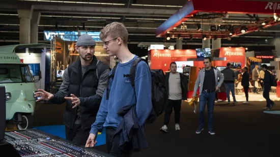 Das Bild zeigt zwei Männer, die auf der Prolight + Sound vor einem Mischpult stehen und darüber reden. Im Hintergrund sind weiere Besucher der Messe zu sehen.