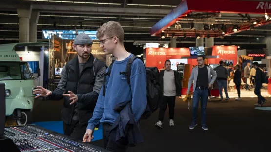 Das Bild zeigt zwei Männer, die auf der Prolight + Sound vor einem Mischpult stehen und darüber reden. Im Hintergrund sind weiere Besucher der Messe zu sehen.