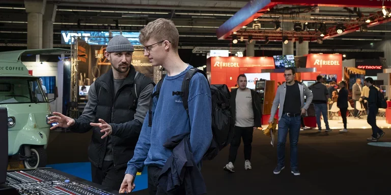Halle 11 auf der Prolight + Sound Das Bild zeigt zwei Männer, die auf der Prolight + Sound vor einem Mischpult stehen und darüber reden. Im Hintergrund sind weiere Besucher der Messe zu sehen.