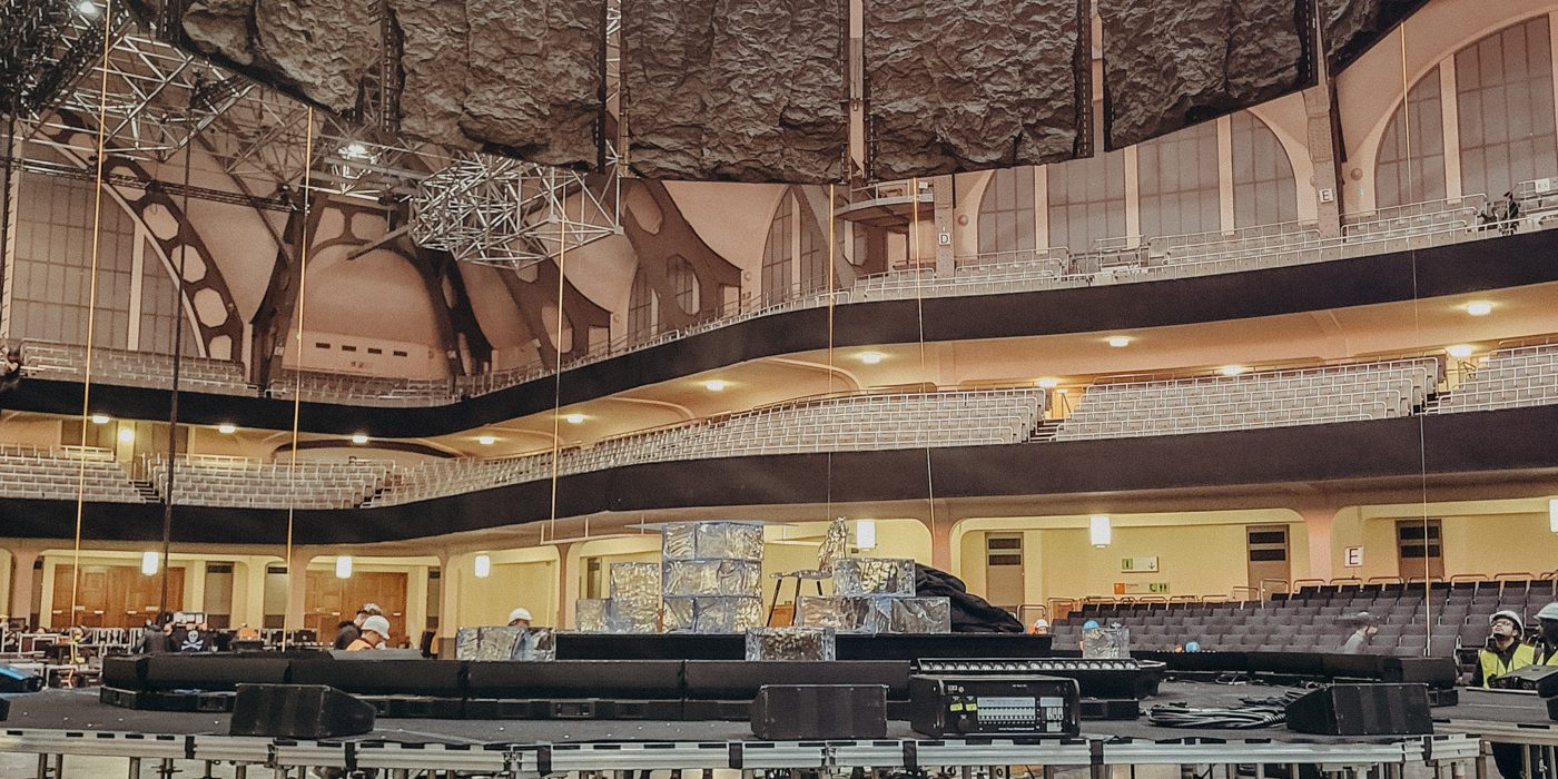 Das Bild zeigt den Aufbau einer Bühne in einer großen Veranstaltungshalle, der Festhalle Frankfurt. Zu sehen ist eine runde Bühnenkonstruktion, die auf zahlreichen höhenverstellbaren Gerüstfüßen mit Rollen montiert ist. Über der Bühne hängen schwere Vorhänge oder akustische Elemente sowie Teile der Licht- und Tontechnik. Im Hintergrund sind die Sitzreihen und die markante Architektur der Halle zu erkennen. Es handelt sich um einen technischen Aufbauprozess, bei dem die Bühne noch nicht vollständig fertiggestellt ist.