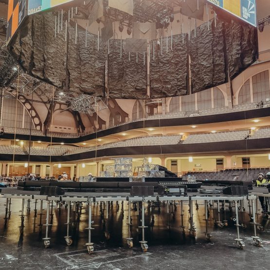 Das Bild zeigt den Aufbau einer Bühne in einer großen Veranstaltungshalle, der Festhalle Frankfurt. Zu sehen ist eine runde Bühnenkonstruktion, die auf zahlreichen höhenverstellbaren Gerüstfüßen mit Rollen montiert ist. Über der Bühne hängen schwere Vorhänge oder akustische Elemente sowie Teile der Licht- und Tontechnik. Im Hintergrund sind die Sitzreihen und die markante Architektur der Halle zu erkennen. Es handelt sich um einen technischen Aufbauprozess, bei dem die Bühne noch nicht vollständig fertiggestellt ist.