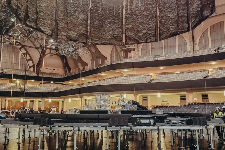 Das Bild zeigt den Aufbau einer Bühne in einer großen Veranstaltungshalle, der Festhalle Frankfurt. Zu sehen ist eine runde Bühnenkonstruktion, die auf zahlreichen höhenverstellbaren Gerüstfüßen mit Rollen montiert ist. Über der Bühne hängen schwere Vorhänge oder akustische Elemente sowie Teile der Licht- und Tontechnik. Im Hintergrund sind die Sitzreihen und die markante Architektur der Halle zu erkennen. Es handelt sich um einen technischen Aufbauprozess, bei dem die Bühne noch nicht vollständig fertiggestellt ist.