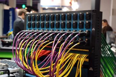 Das Bild zeigt ein professionelles Kabelpatchfeld, das auf einer Messe ausgestellt wird. Das Patchfeld verfügt über mehrere Reihen von XLR-Anschlüssen, die in einem strukturierten Raster angeordnet sind. Zahlreiche Kabel in unterschiedlichen Farben, darunter Gelb, Lila und Rot, sind mit den Anschlüssen verbunden. Diese farbliche Codierung deutet auf eine systematische Organisation hin, um verschiedene Signalwege zu kennzeichnen. Im Hintergrund erkennt man die Messeatmosphäre, mit Personen und weiteren technischen Ausstellungsstücken. Das Bild vermittelt einen Eindruck von der Komplexität und Präzision, die bei der Signalverarbeitung und -verteilung in der Veranstaltungstechnik erforderlich ist.