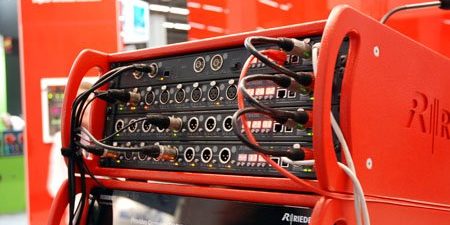 Das Bild zeigt eine technische Audiokonfiguration auf einer Messe. Im Vordergrund steht ein leuchtend rotes Rack mit zahlreichen Audiogeräten, das mit verschiedenen Kabeln verbunden ist. Diese Geräte, vermutlich Audio-Interfaces oder Signalprozessoren, verfügen über zahlreiche Eingangs- und Ausgangsanschlüsse. Die Kabel sind sorgfältig angeschlossen, was auf eine Demonstration oder Testeinrichtung hinweist. Im Hintergrund ist die Messeumgebung zu erkennen, die durch rote Wände und Informationsschilder geprägt ist. Das Logo des Herstellers „Riedel“ ist gut sichtbar und deutet auf professionelle Audiotechnologie hin. Die Szene vermittelt einen Eindruck von moderner Technik und zeigt ein Beispiel für Audio-Netzwerkintegration im professionellen Bereich.