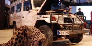 Das Bild zeigt eine Ausstellungsszene, in der ein Allradfahrzeug vom Typ Toyota Land Cruiser prominent im Vordergrund steht. Das Fahrzeug ist mit Expeditionsausrüstung beladen, einschließlich Gepäck und zusätzlicher Ausrüstung auf dem Dachträger. Die Motorhaube ist geöffnet, was möglicherweise technische Details des Fahrzeugs präsentiert. Links im Bild ist ein ausgestopfter Leopard abgebildet, dessen Maul geöffnet ist, als ob er faucht. Im Hintergrund ist eine Messeatmosphäre zu erkennen, mit großen Bildschirmen und weiteren Ausstellungsbereichen. Die Szene vermittelt eine Mischung aus Abenteuer- und Präsentationsthema, das vermutlich auf Expeditionstechnologie oder Erlebnisreisen hinweist.