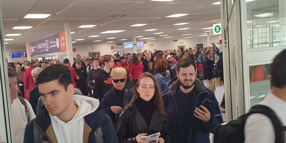 Auf dem Bild ist ein stark frequentierter Eingangsbereich einer Messe oder Veranstaltungshalle zu sehen – offenbar gerade zur Öffnung oder unmittelbar nach Beginn des Events. Hunderte Menschen strömen durch breite Türen in die Halle, viele mit Tickets, Smartphones oder Broschüren in der Hand. Die Stimmung wirkt geschäftig, aber geordnet. Im Hintergrund erkennt man ein großes Banner mit dem Schriftzug "Production + DJ" sowie Hallennummern (z. B. 12.0, F 46), was auf einen Messekontext hindeutet – vermutlich die Prolight + Sound 2025. Auch die Wegweiser und Monitore zur Orientierung sowie die „S“-Schilder (vielleicht für „Service“ oder „Shuttle“) verstärken den Eindruck eines professionell organisierten Großevents. Das Bild vermittelt den Moment des Ansturms: viele Besucher, teils in Gruppen, teils einzeln, die sich auf den Weg in die Ausstellungsbereiche machen. Es zeigt, wie groß das Interesse an der Veranstaltung ist.