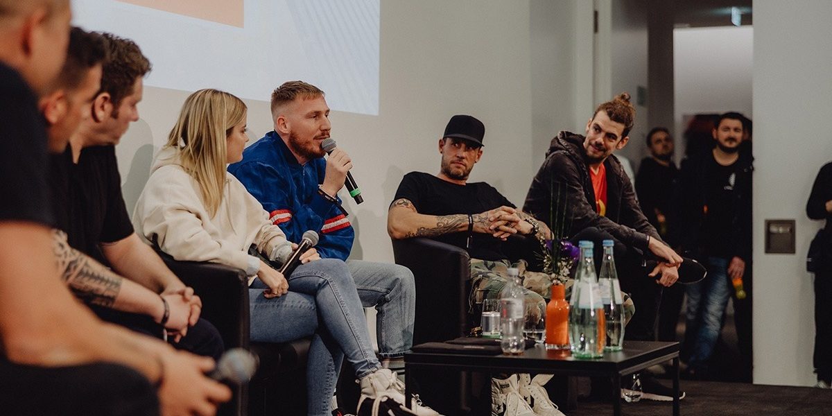 MIXCON x BVD present DJ- & Producer Conference Das Bild zeigt eine Podiumsdiskussion auf der MIXCON x BVD DJ- & Producer Conference. Mehrere Personen sitzen auf einer Bühne in einem modernen Konferenzraum. Eine Person in einer blauen Jacke mit roten Streifen spricht in ein Mikrofon, während die anderen Panelisten aufmerksam zuhören. Die Atmosphäre wirkt entspannt und professionell. Im Vordergrund ist ein Tisch mit Wasserflaschen und Blumen zu sehen. Im Hintergrund stehen einige Zuschauer, die das Gespräch verfolgen. Das MIXCON-Logo in der unteren rechten Ecke des Bildes weist auf den Veranstaltungszusammenhang hin.