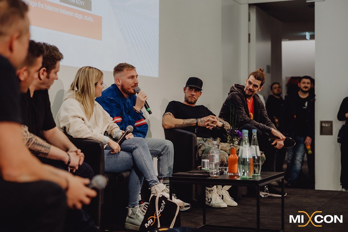 Das Bild zeigt eine Podiumsdiskussion auf der MIXCON x BVD DJ- & Producer Conference. Mehrere Personen sitzen auf einer Bühne in einem modernen Konferenzraum. Eine Person in einer blauen Jacke mit roten Streifen spricht in ein Mikrofon, während die anderen Panelisten aufmerksam zuhören. Die Atmosphäre wirkt entspannt und professionell. Im Vordergrund ist ein Tisch mit Wasserflaschen und Blumen zu sehen. Im Hintergrund stehen einige Zuschauer, die das Gespräch verfolgen. Das MIXCON-Logo in der unteren rechten Ecke des Bildes weist auf den Veranstaltungszusammenhang hin.
