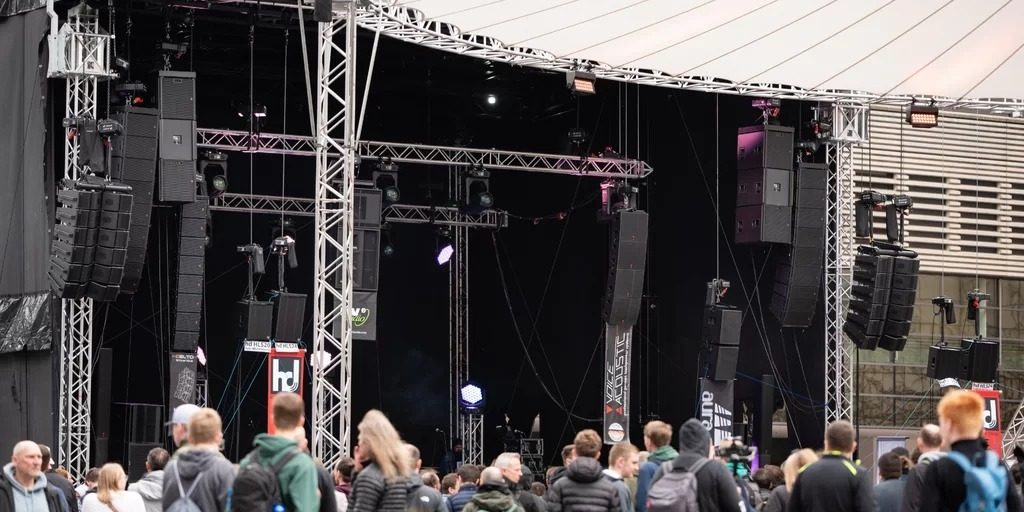 ProAudio-Angebot Live Sound Arena Das Bild zeigt eine große Outdoor-Bühne mit einer professionellen PA-Beschallungsanlage auf der Prolight + Sound Messe. Die Bühne ist von einem stabilen Traversensystem umgeben, an dem mehrere Line-Array-Lautsprechersysteme von verschiedenen Herstellern hängen. Im Vordergrund befindet sich eine Menschenmenge, die sich die Präsentationen anhört oder auf die nächsten Vorführungen wartet. Die Atmosphäre wirkt dynamisch und zeigt, wie die Live Sound Arena als zentrale Plattform für Audiotechnik-Enthusiasten und Fachbesucher dient. Die Logos der teilnehmenden Hersteller wie „hd“ (Harmonic Design) und „Voice-Acoustic“ sind an den Lautsprechersystemen sichtbar, was darauf hinweist, dass hier neueste Beschallungstechnologien präsentiert werden.
