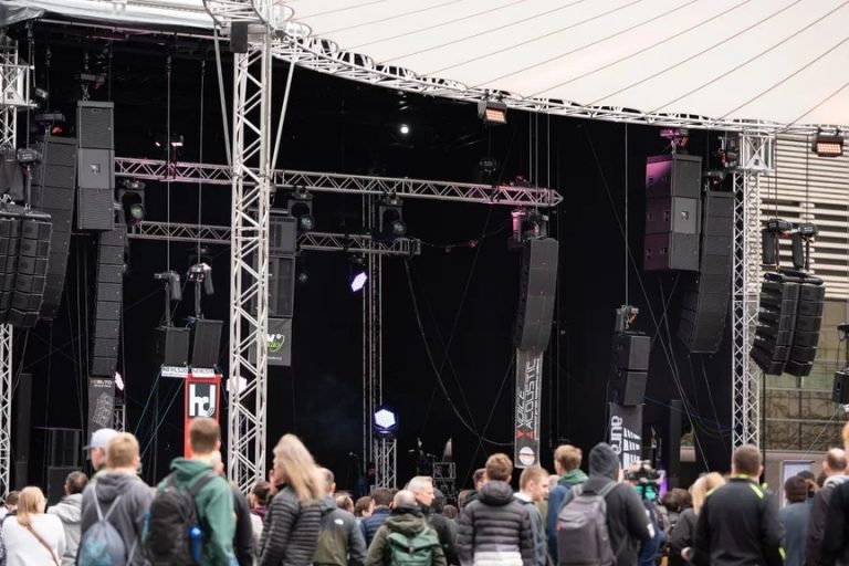 Das Bild zeigt eine große Outdoor-Bühne mit einer professionellen PA-Beschallungsanlage auf der Prolight + Sound Messe. Die Bühne ist von einem stabilen Traversensystem umgeben, an dem mehrere Line-Array-Lautsprechersysteme von verschiedenen Herstellern hängen. Im Vordergrund befindet sich eine Menschenmenge, die sich die Präsentationen anhört oder auf die nächsten Vorführungen wartet. Die Atmosphäre wirkt dynamisch und zeigt, wie die Live Sound Arena als zentrale Plattform für Audiotechnik-Enthusiasten und Fachbesucher dient. Die Logos der teilnehmenden Hersteller wie „hd“ (Harmonic Design) und „Voice-Acoustic“ sind an den Lautsprechersystemen sichtbar, was darauf hinweist, dass hier neueste Beschallungstechnologien präsentiert werden.
