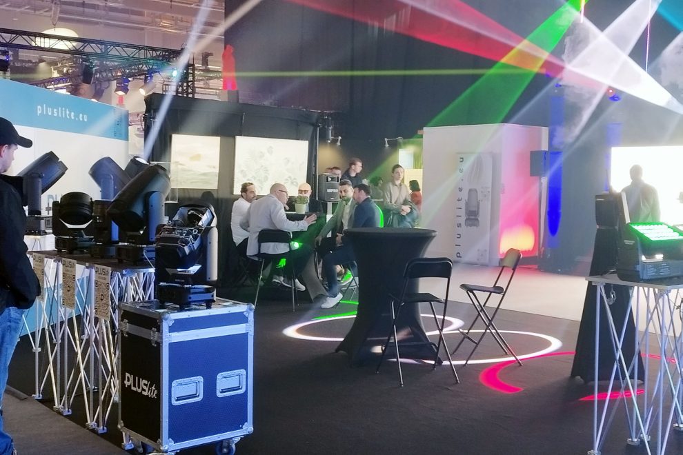 Messestand von Pluslite mit Scheinwerfern, Lichtstrahlen und Besuchergruppe in Gesprächen.