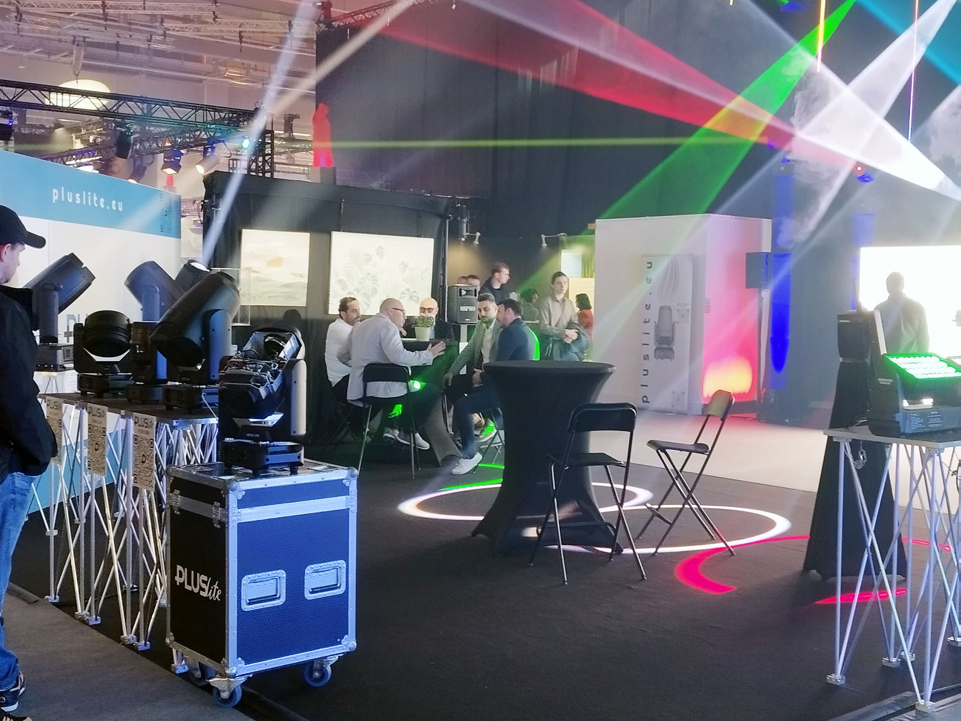 Messestand von Pluslite mit Scheinwerfern, Lichtstrahlen und Besuchergruppe in Gesprächen.