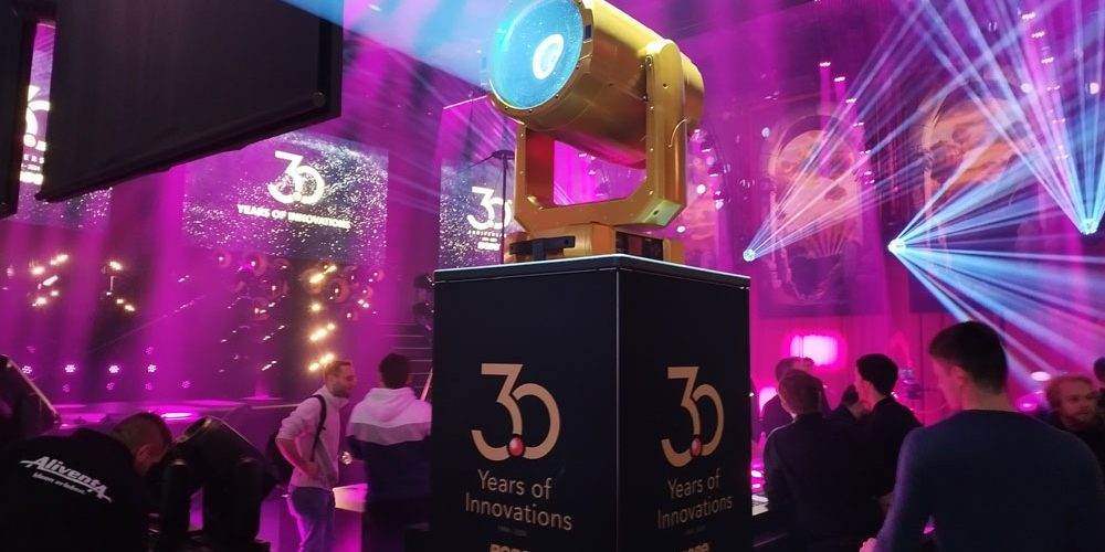 Robe Lighting Show Prolight + Sound 2024 Das Bild zeigt eine Bühne mit einer beeindruckenden Lichtshow, die von einem zentral positionierten, großen Scheinwerfer dominiert wird. Der Scheinwerfer steht auf einem Podest mit der Aufschrift „30 Years of Innovations – ROBE“ und wird durch eine dynamische Lichtinszenierung in Szene gesetzt. Im Hintergrund sind farbige Lichtstrahlen und projizierte Logos sichtbar, die die Atmosphäre einer professionellen Veranstaltung oder Jubiläumsfeier verstärken. Menschen im Vordergrund und in der Umgebung deuten auf eine Messe oder Präsentation hin, bei der Lichttechnik und Innovationen im Mittelpunkt stehen. Die Umgebung ist in lebendigen Pink- und Blautönen beleuchtet, was der Szenerie eine futuristische und festliche Stimmung verleiht.
