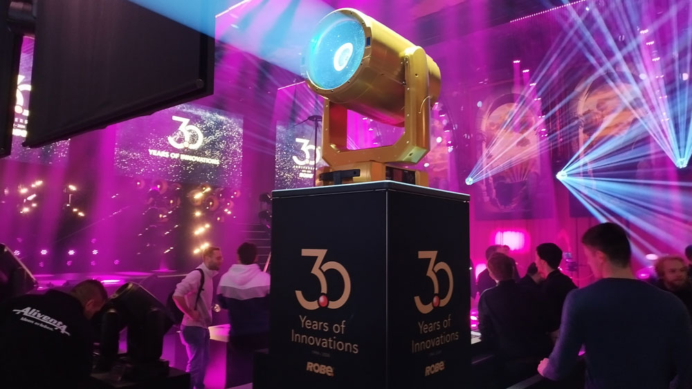 Das Bild zeigt eine Bühne mit einer beeindruckenden Lichtshow, die von einem zentral positionierten, großen Scheinwerfer dominiert wird. Der Scheinwerfer steht auf einem Podest mit der Aufschrift „30 Years of Innovations – ROBE“ und wird durch eine dynamische Lichtinszenierung in Szene gesetzt. Im Hintergrund sind farbige Lichtstrahlen und projizierte Logos sichtbar, die die Atmosphäre einer professionellen Veranstaltung oder Jubiläumsfeier verstärken. Menschen im Vordergrund und in der Umgebung deuten auf eine Messe oder Präsentation hin, bei der Lichttechnik und Innovationen im Mittelpunkt stehen. Die Umgebung ist in lebendigen Pink- und Blautönen beleuchtet, was der Szenerie eine futuristische und festliche Stimmung verleiht.