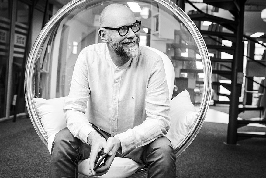 Ein lächelnder Mann mit Brille sitzt entspannt in einem modernen runden Stuhl.