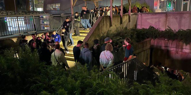 Das Bild zeigt eine größere Menschenmenge, die sich auf einer Treppe und in einem Eingangsbereich versammelt hat. Die Szenerie spielt sich nachts ab, und die Umgebung ist durch künstliches Licht beleuchtet. Die Personen wirken entspannt, einige unterhalten sich, während andere sich umsehen oder auf den Eingang zusteuern. Die Architektur deutet auf eine urbane Umgebung hin, mit Betonwänden, Geländern und einer überdachten Eingangsstruktur. Im Hintergrund sind moderne Gebäude und Lichter zu sehen, die eine lebendige Nachtatmosphäre schaffen. Möglicherweise handelt es sich um den Einlass zu einer Veranstaltung oder einem Club.