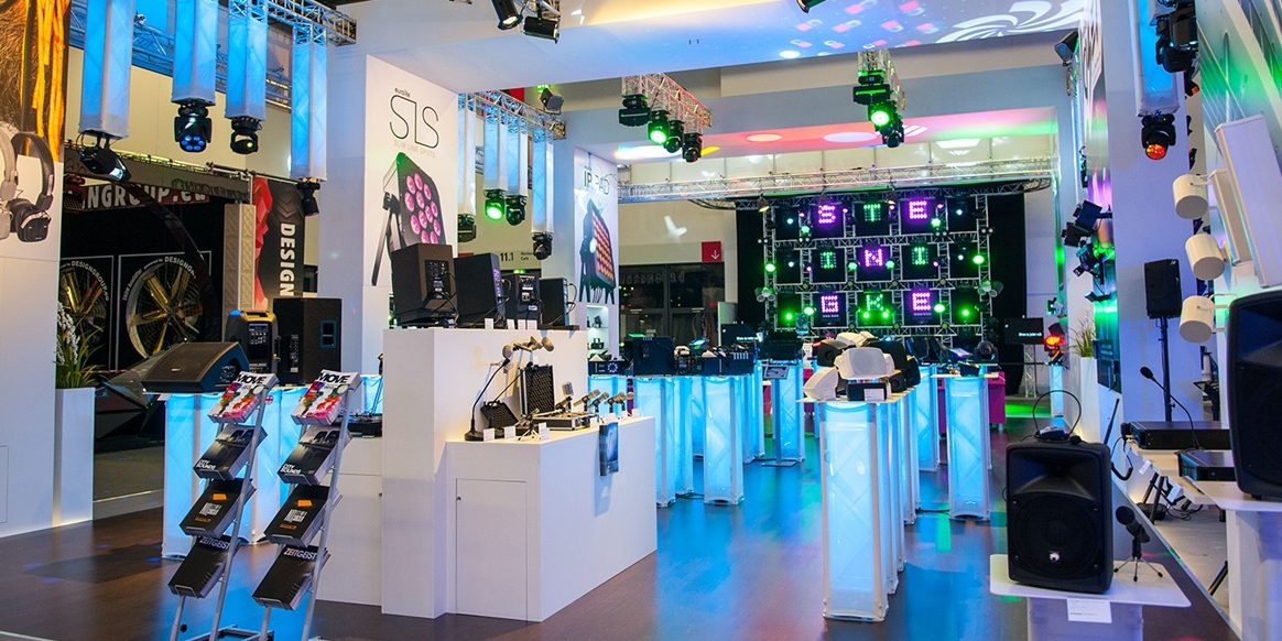 steinigke_pls_2015 Das Bild zeigt einen farbenfrohen Messestand auf einer Veranstaltung, vermutlich der Prolight + Sound 2015. Der Stand ist von Steinigke Showtechnic und präsentiert verschiedene Beleuchtungs- und Audio-Produkte. Im Vordergrund sind auf beleuchteten Podesten Mikrofone, Lautsprecher und andere technische Geräte ausgestellt. Im Hintergrund dominiert eine beeindruckende Lichtinstallation mit leuchtenden Buchstaben, die den Stand optisch aufwertet. Über dem gesamten Bereich hängen bunte Scheinwerfer und Leuchten, die ein lebendiges, professionelles Ambiente schaffen. Der Stand ist hell und einladend gestaltet, mit modernen Designelementen und einem klaren Fokus auf die Präsentation von Veranstaltungstechnik.
