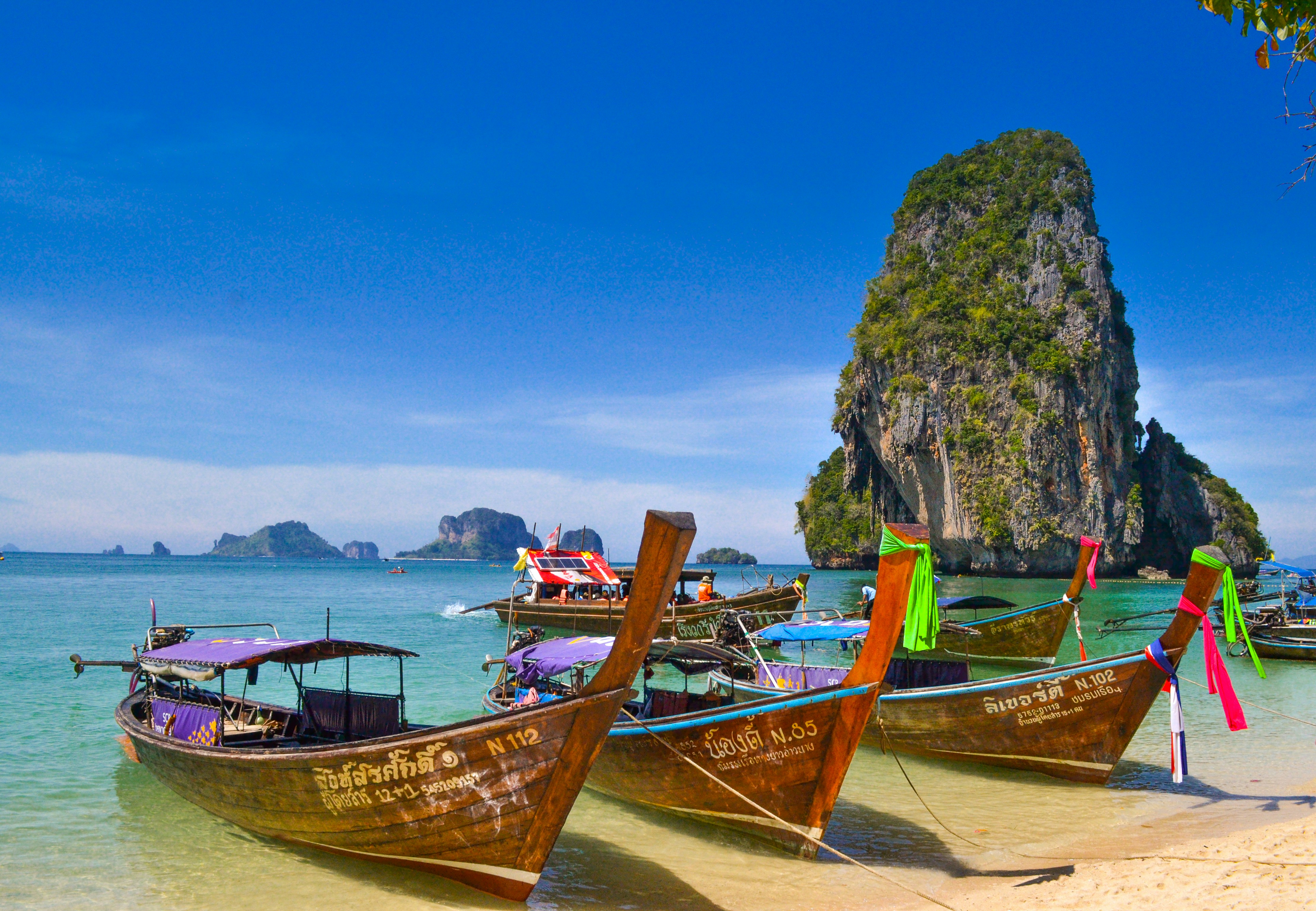 Longtail-Boote an einem tropischen Strand in Thailand mit Felseninsel im Meer.