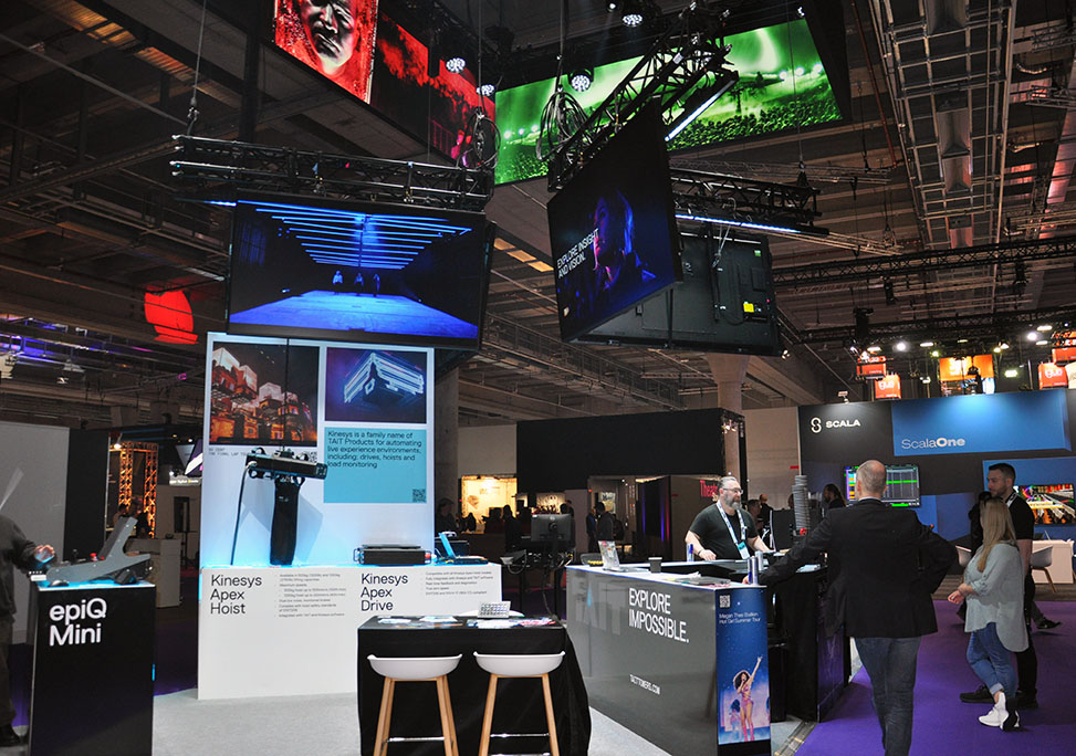 Messestand von Tait mit epiQ Mini Console, Bildschirmen und Besucherinnen und Besuchern auf Messe.