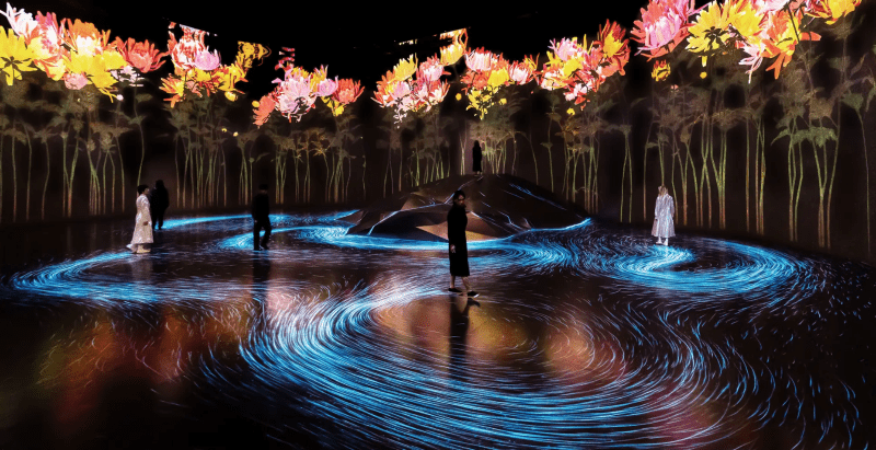 Immersive space with glowing flowers and flowing projections, people moving within it.