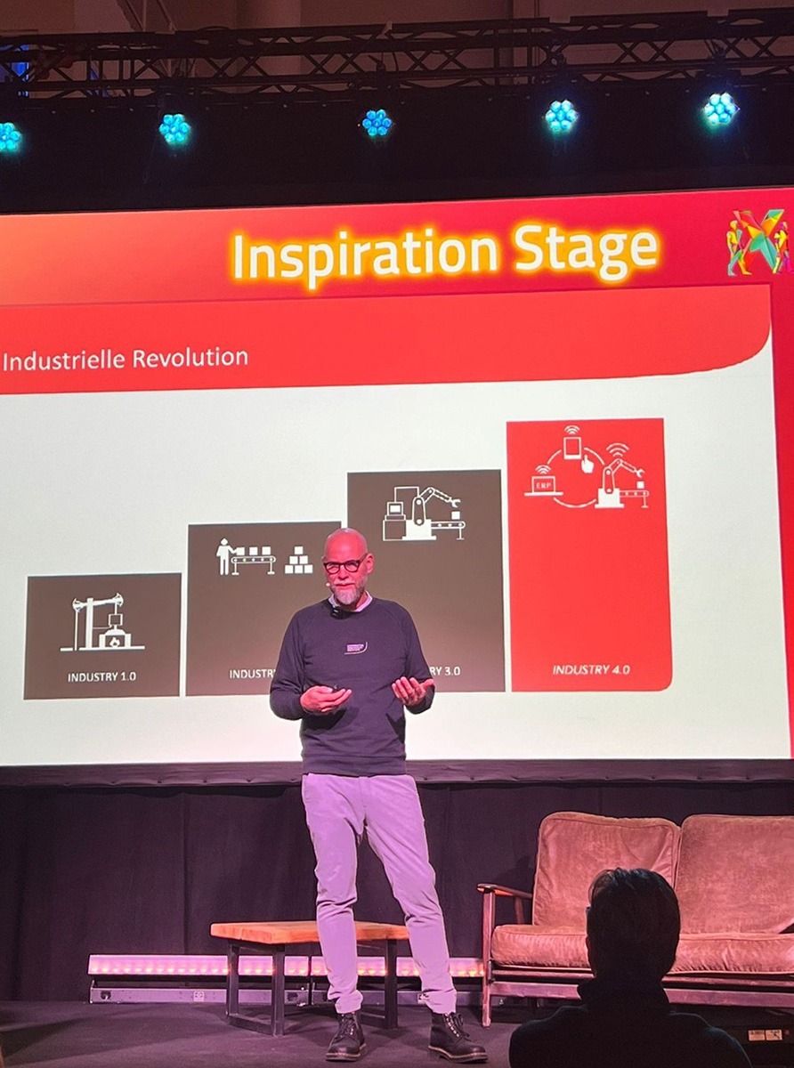 Ein Redner steht auf der Bühne vor einer Präsentation zur industriellen Revolution.