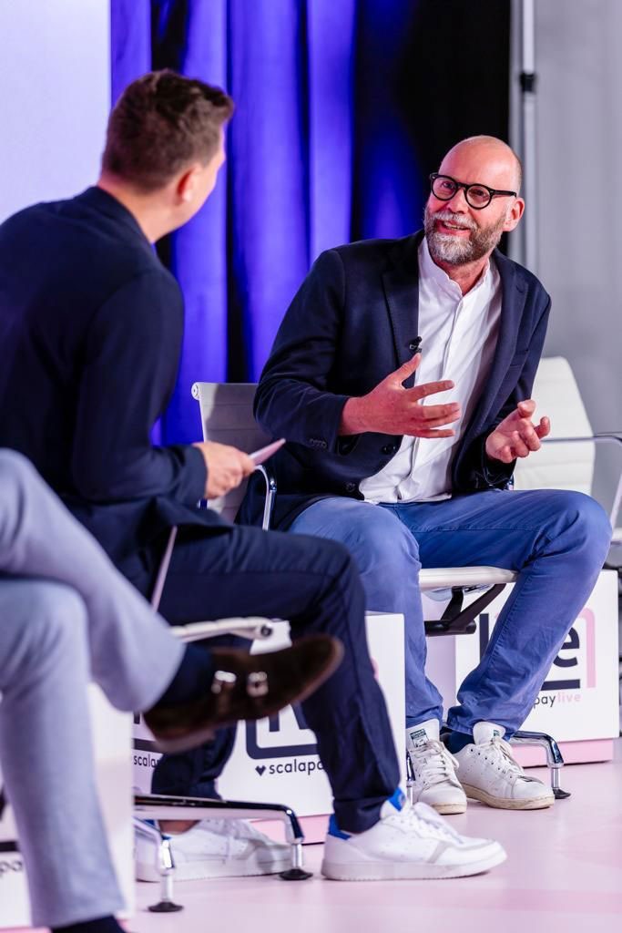 Zwei Männer sitzen auf einer Bühne und sprechen angeregt in einem Interviewgespräch.