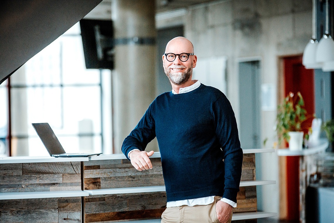 Ein lächelnder Mann mit Brille steht entspannt an einem Tresen in einem modernen Büro.