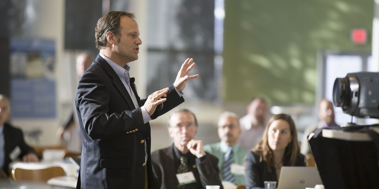 A3E Konferenz Das Bild zeigt eine Szene, in der ein Mann in einem dunklen Anzug eine Präsentation oder einen Vortrag hält. Er steht im Vordergrund und gestikuliert mit den Händen, während er spricht. An seinem Revers ist ein Mikrofon befestigt, was auf eine professionelle Veranstaltung hindeutet. Im Hintergrund sitzen mehrere Personen an runden Tischen und hören aufmerksam zu. Einige Teilnehmer haben Notebooks oder Unterlagen vor sich, was darauf hinweist, dass es sich um ein Meeting, eine Konferenz oder ein Seminar handelt. Auf der rechten Seite ist eine Kamera zu sehen, die möglicherweise den Vortrag aufzeichnet. Die Atmosphäre wirkt geschäftlich und professionell.